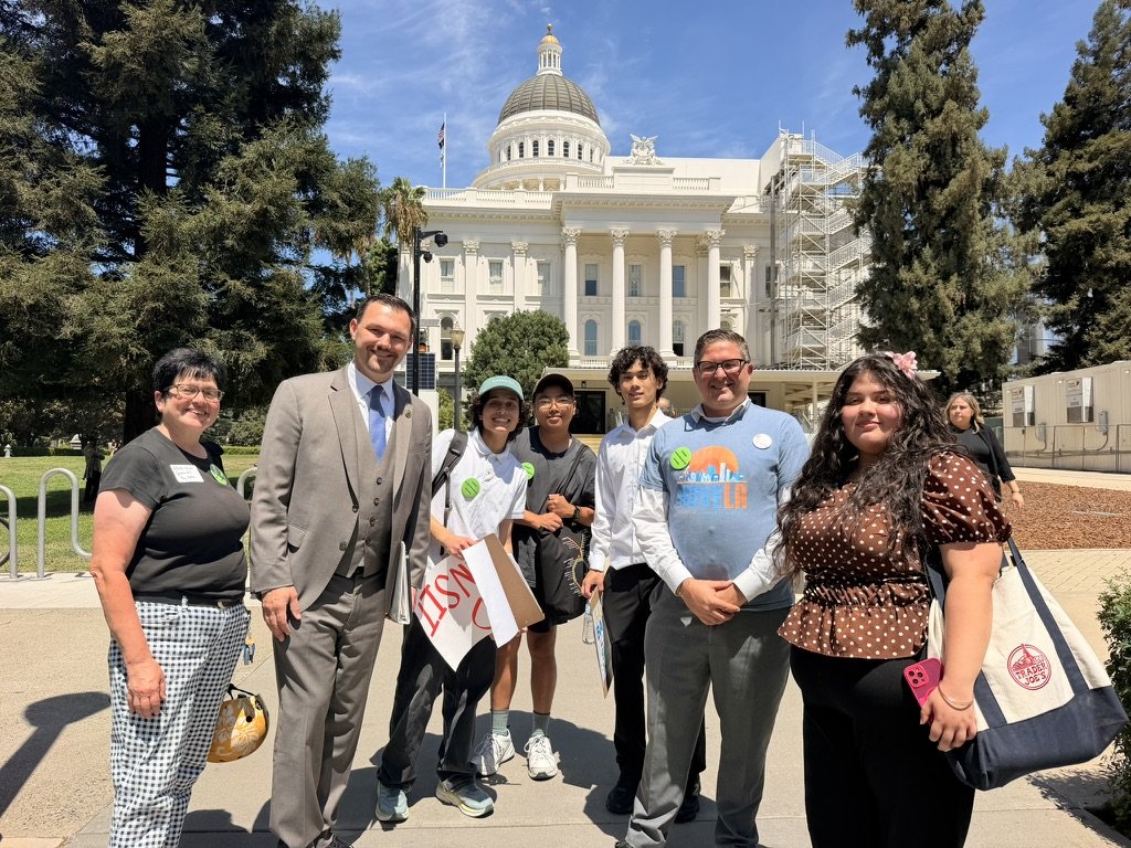 seven people standing in front of the California Capitol building, white facade with four columns and a domed peak. The people holding signs and wearing matching green pins.